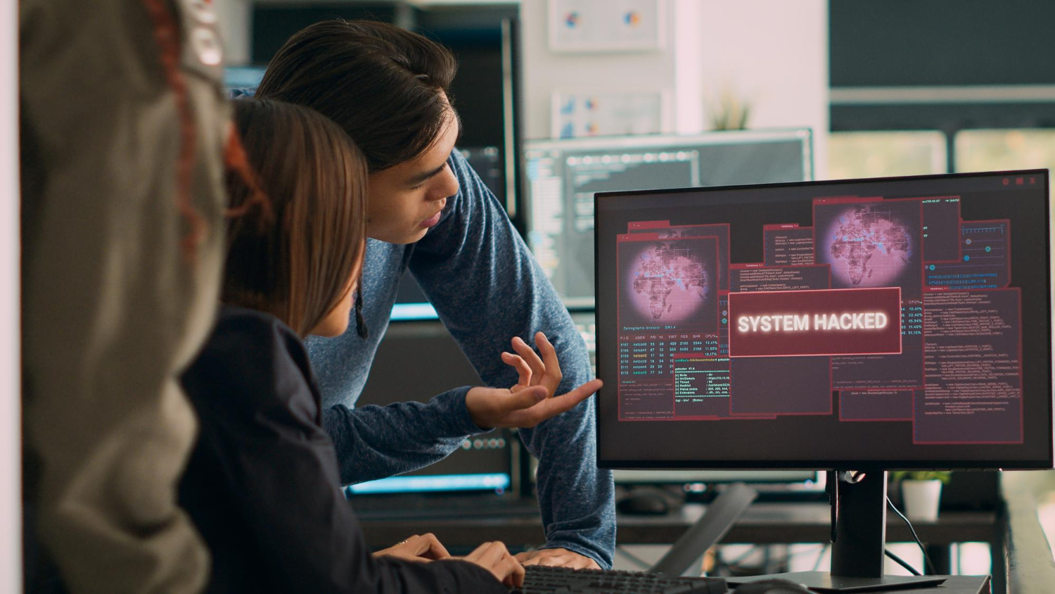 The image shows Two individuals observing a computer screen displaying a virus, indicating a system hack. 