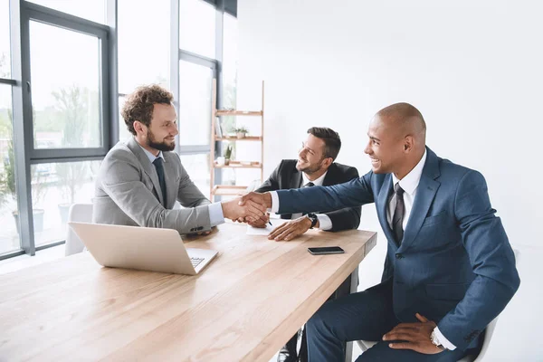 The image shows Three business professionals shaking hands during a meeting. 